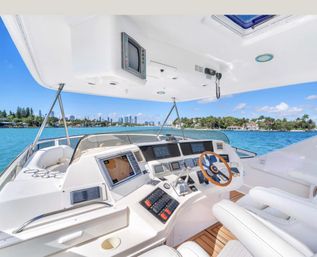 Luxury yacht helm on a sunny day — wooden steering wheel, multi-screen navigation console and white leather seats overlooking turquoise water and a palm-lined coastal skyline.