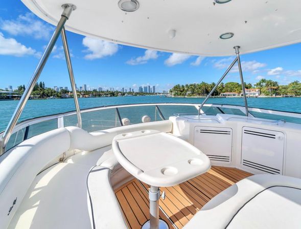 View from a luxury yacht's sun deck with white cushioned seating and table, turquoise bay, palm-lined shoreline and distant Miami skyline under a bright blue sky