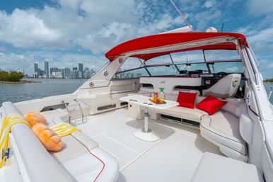 White luxury motorboat lounge with red canopy, bright pillows and a table with drinks, anchored on a blue bay with a distant waterfront city skyline under a partly cloudy sky