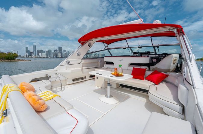 White luxury motorboat lounge with red canopy, bright pillows and a table with drinks, anchored on a blue bay with a distant waterfront city skyline under a partly cloudy sky