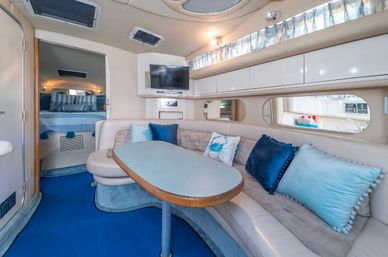 Cozy yacht cabin salon with curved cream leather dinette and oval table, navy and aqua throw pillows, wall-mounted TV, small galley visible through a side window, and a forward sleeping berth with striped bedding.
