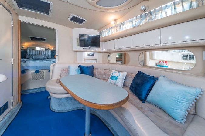 Cozy yacht cabin salon with curved cream leather dinette and oval table, navy and aqua throw pillows, wall-mounted TV, small galley visible through a side window, and a forward sleeping berth with striped bedding.