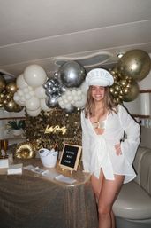 Smiling bride-to-be wearing a white 'BRIDE' cap and cover-up poses beside a table decorated with gold and silver balloons, a sequin backdrop, champagne in an ice bucket, and a small letter board at a bachelorette-style indoor celebration.