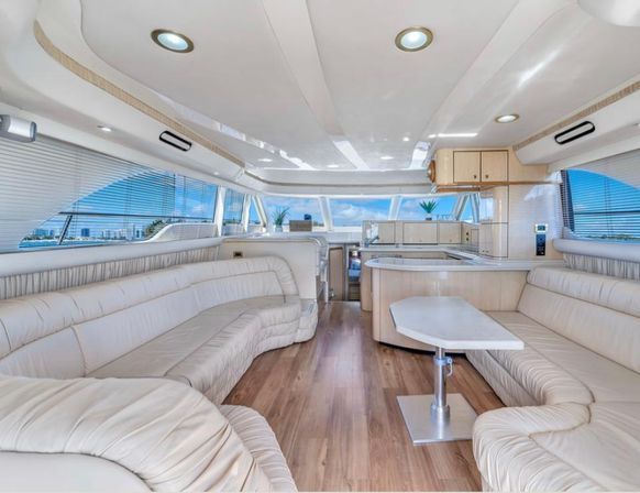 Sunlit luxury yacht salon with cream leather wraparound seating, wood-look flooring, small white table and open galley, panoramic windows framing blue water and a coastal skyline.