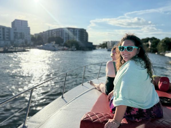 Smiling woman in turquoise sunglasses wrapped in a towel relaxing on a boat bow near an urban waterfront marina at golden hour, sun sparkling on the water and apartment buildings in the background.