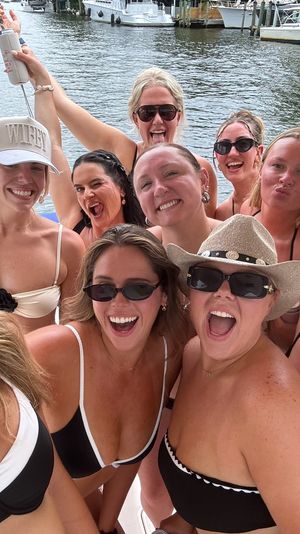 Lively group of women in swimsuits and sunglasses taking a selfie on a boat at a marina, smiling and cheering with docks and yachts in the background.
