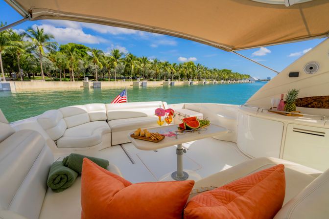 Luxury yacht aft deck with curved white leather seating, small table with tropical cocktails, watermelon and snacks, orange pillows and rolled towels, American flag and palm‑lined turquoise waterfront under a blue sky