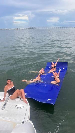 Group of friends in swimsuits laughing and splashing on a large blue floating mat tethered to a white boat in a calm coastal bay with a distant bridge and cloudy sky