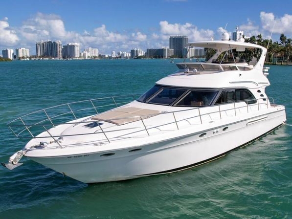 White luxury motor yacht cruising turquoise waters with a coastal city skyline and palm trees under a sunny blue sky