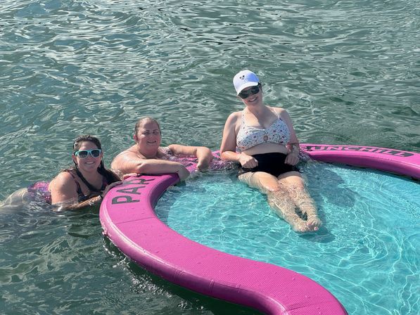 Three people relaxing on a bright pink circular floating lounge in clear lake water on a sunny summer day, wearing swimsuits, sunglasses and a white cap.