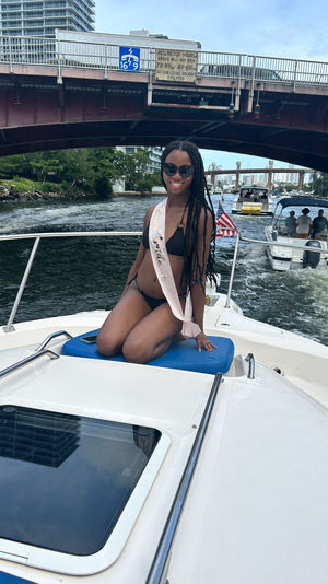 Smiling woman in a black bikini wearing a 'Bride to Be' sash kneels on the bow of a motorboat cruising an urban waterway under a low bridge, with an American flag, other boats, and city buildings in the background