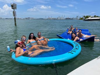 Group of friends in swimsuits and heart-shaped sunglasses lounging and cheering with canned drinks on a round blue floating pool and linked mats in a sunny harbor, with yachts, cargo cranes, and city skyline on the horizon.