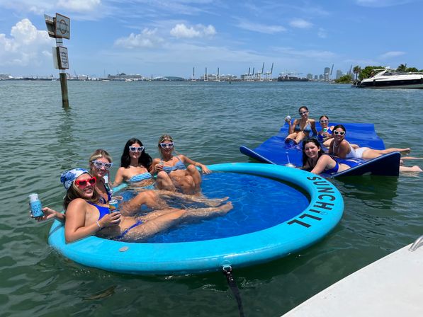 Group of friends in swimsuits and heart-shaped sunglasses lounging and cheering with canned drinks on a round blue floating pool and linked mats in a sunny harbor, with yachts, cargo cranes, and city skyline on the horizon.