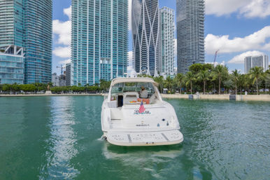 Sleek white motor yacht with an American flag on turquoise water in front of a palm-lined Miami Beach waterfront and tall glass high-rise skyline under a blue sky.