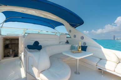 Sun-soaked luxury yacht seating with white curved leather sofa, blue bimini canopy, table with champagne and flutes, turquoise sea and distant coastal skyline.