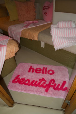 Plush pink floor mat reading 'hello beautiful' in a cozy pink-themed sleeping nook with striped towels, folded blankets, a heart 'Cutie Pie' pillow and floral accent cushions.