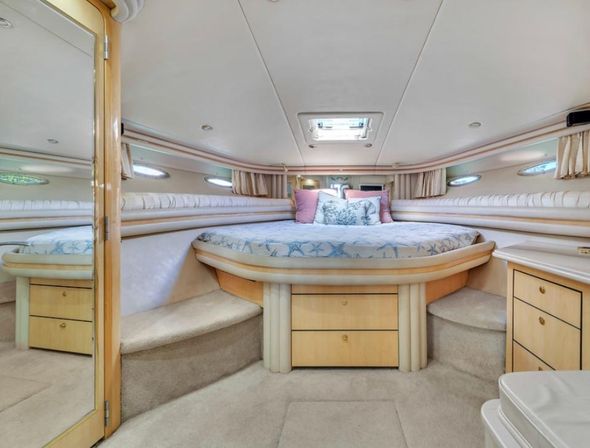 Cozy luxury yacht cabin interior with a raised V-shaped bed, light wood cabinets and steps, oval porthole windows, overhead skylight, and pastel pillows on sea‑themed bedding.
