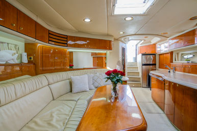 Luxury yacht salon with curved cream leather seating, glossy cherry wood cabinets and table topped with a vase of red roses, compact galley with sink and fridge, skylight and stairs to deck