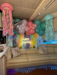 Under-the-sea themed indoor birthday lounge with pastel balloon garland, metallic accents and hanging jellyfish paper lanterns, rainbow tassel backdrop and neon "Happy Birthday" sign above a beige sofa with turtle throw pillows.