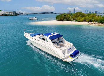 Sunlit white motor yacht with blue canvas top cruising turquoise water past a small sandy island and mangroves, with a bridge and city skyline on the horizon.