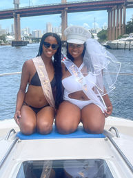 Bachelorette boat party on an urban waterfront: two smiling friends in swimsuits, bride-to-be wearing veil, 'BRIDE' hat and sash, city skyline and bridge in the background.