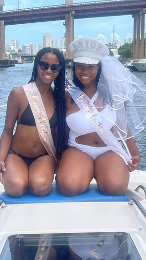 Two smiling women in swimsuits wearing 'Bride to Be' sash and bridal hat with veil and 'Team Bride' sash, posing on a boat during a bachelorette party with an urban skyline, bridge, and marina in the background.