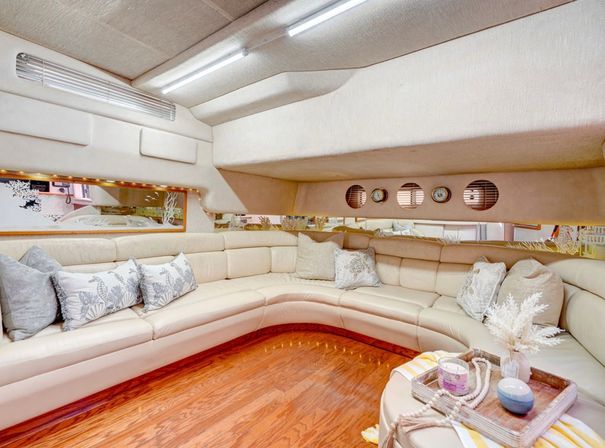 Cozy luxury yacht salon with cream leather U-shaped sofa, decorative pillows, warm wood flooring, porthole windows and a tray of coastal decor.