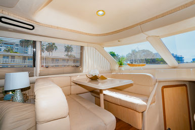Bright yacht interior dining nook with cream leather wraparound seating, small wood table, decorative model ship and potted plant, windows with blinds revealing palm trees and a marina view.