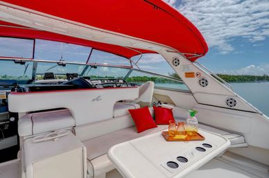 Luxury white motor yacht seating area under a red canopy with red throw pillows and glassware on a table, overlooking calm turquoise coastal waters and blue sky