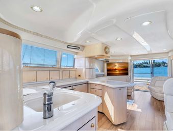 Bright luxury yacht interior — open-plan cream galley and salon with curved white countertops, stainless faucet, hardwood floors, comfortable seating and large windows showing blue water and marina shoreline.