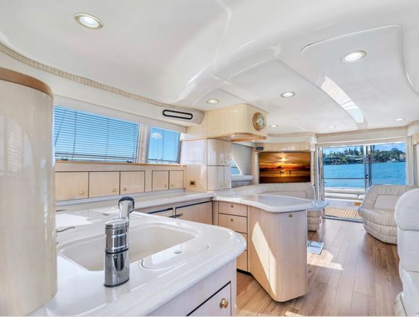 Bright luxury yacht interior — open-plan cream galley and salon with curved white countertops, stainless faucet, hardwood floors, comfortable seating and large windows showing blue water and marina shoreline.