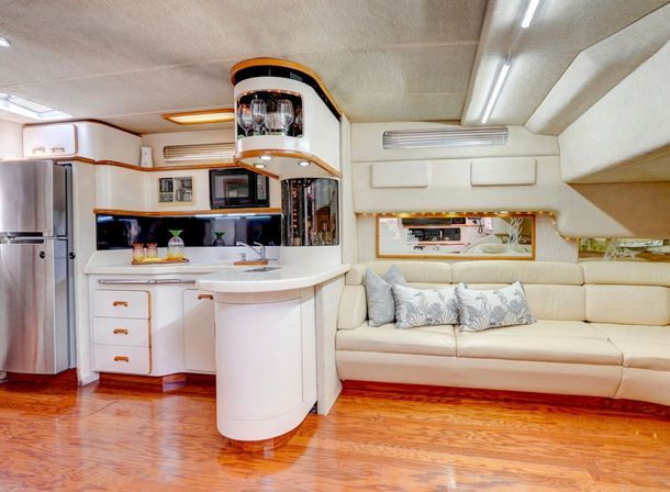 Modern luxury yacht interior with cream leather salon and decorative pillows, glossy wood floors, curved white galley countertop with sink, stainless steel refrigerator, glassware display and ambient LED lighting.