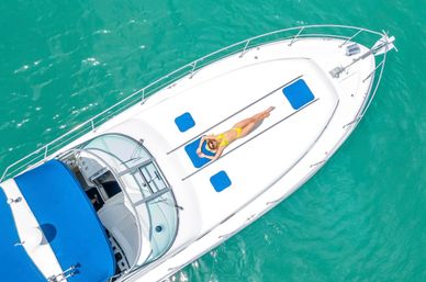 Aerial view of a white motor yacht on clear turquoise water with a person in a yellow swimsuit relaxing on blue deck pads, sunbathing and enjoying the tropical scene.