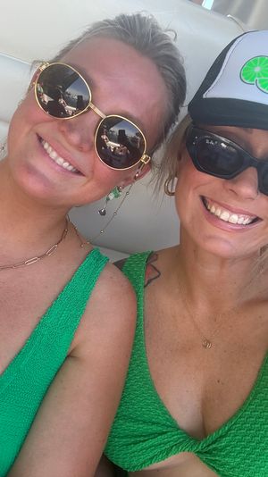 Sunny boat selfie of two smiling friends in matching green swimsuits, sunglasses and a lime‑graphic trucker hat