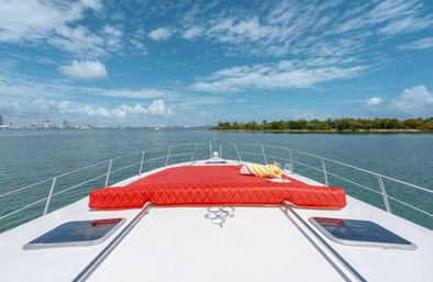 Bow of a white yacht with a bright red quilted sunpad, towel and drinks cruising a calm turquoise bay past a tree-lined island and distant city skyline under a blue sky with scattered clouds.