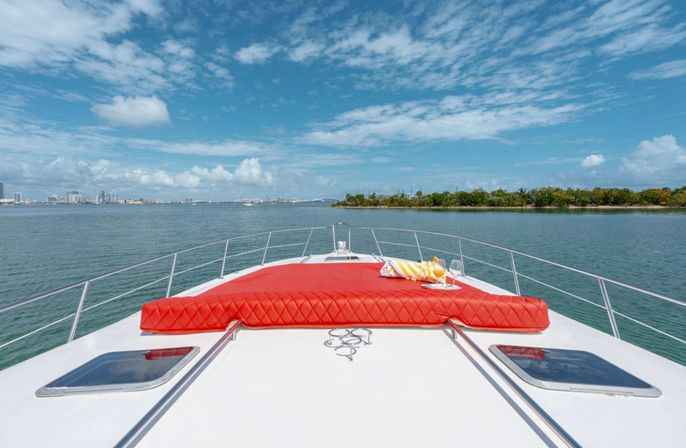 Bow of a white yacht with a bright red quilted sunpad, towel and drinks cruising a calm turquoise bay past a tree-lined island and distant city skyline under a blue sky with scattered clouds.