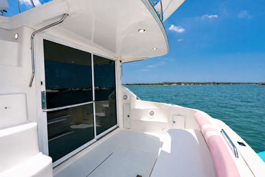 Sunlit yacht stern deck with sliding glass doors, white built-in seating and stainless handrails overlooking turquoise coastal waters and a clear blue sky.