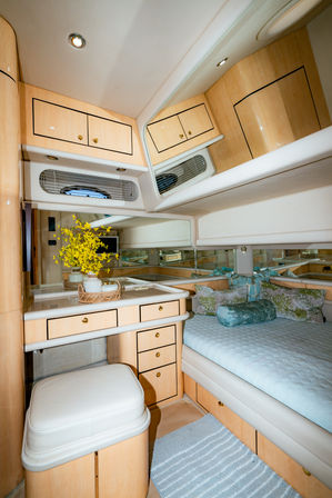 Cozy luxury yacht cabin interior with light wood cabinetry, mirrored vanity and drawers, upholstered stool, blue quilted bed with decorative pillows and a vase of yellow flowers.