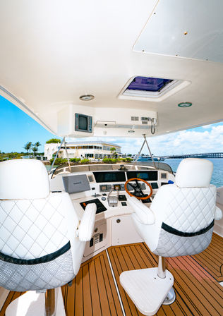 Luxury yacht helm with quilted white captain chairs, wood-trim steering wheel, navigation displays and teak deck overlooking a sunny coastal marina and bridge