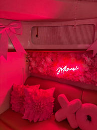 Pink-lit lounge nook with neon "Miami" sign over a flower-covered wall, oversized floral pillows, plush XO cushions and a decorative bow