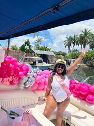 Smiling woman in a captain's hat and white swimsuit wearing a pink bachelorette sash, waving aboard a boat decorated with bright pink and silver balloons, a champagne bucket and palm trees along a sunny tropical waterfront.