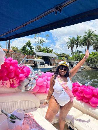 Smiling woman in a captain's hat and white swimsuit wearing a pink bachelorette sash, waving aboard a boat decorated with bright pink and silver balloons, a champagne bucket and palm trees along a sunny tropical waterfront.