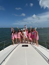 Group of women in pink outfits celebrating a bachelorette party on the bow of a white yacht, coastal waters and boats under a bright blue sky.