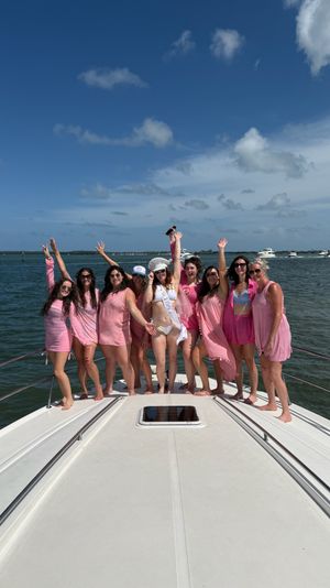 Laughing women in pink beachwear and a bride-to-be in a white sash posing on the bow of a yacht in sunny coastal waters, with boats and a distant bridge under a blue sky