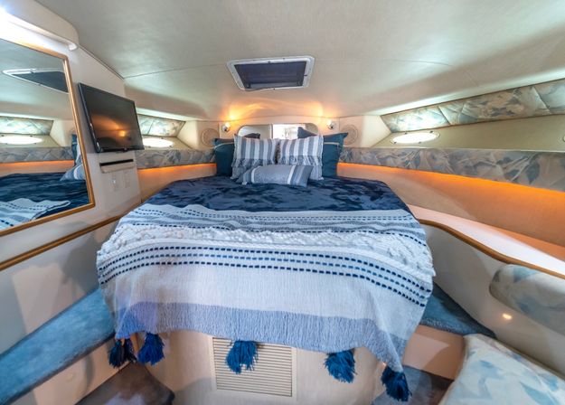 Cozy yacht V-berth cabin with a neatly made bed draped in a blue-striped throw and tassels, striped pillows, porthole windows, wall-mounted TV and warm LED accent lighting.