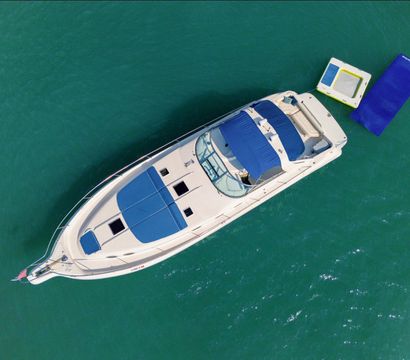 Aerial view of a white luxury motor yacht with blue sun pads and canopy anchored in clear turquoise water beside a blue-and-white floating mat.