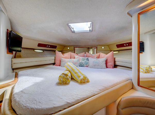 Cozy yacht v-berth cabin interior with quilted bed, coral-themed throw pillows, rolled yellow-striped towels and overhead hatch skylight.