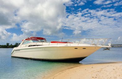 White luxury motor yacht with a red canvas top beached on a sandy tropical shore in turquoise shallow water under a bright blue sky with puffy clouds.