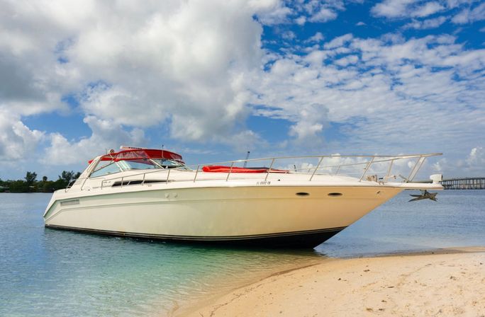 White luxury motor yacht with a red canvas top beached on a sandy tropical shore in turquoise shallow water under a bright blue sky with puffy clouds.