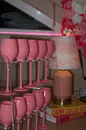 Glossy pink wine glasses on a clear acrylic rack beside a pink patterned lamp, neon pink accent lighting and faux floral garland in a modern pink-themed bar or vanity display